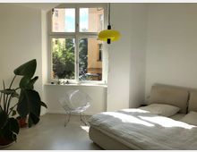 Helles Schlafzimmer mit einem großen Fenster, durch das Tageslicht einfällt; links im Raum steht eine große Zimmerpflanze in einem Topf, ein moderner Drahtstuhl, und rechts ein Bett mit weißer Bettwäsche. Eine gelbe Hängelampe hängt von der Decke herab.