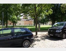 Grüne Parkanlage mit Rasenfläche im Vordergrund, umgeben von Bäumen. Im Hintergrund stehen farbige historische Gebäude, davor sind parkende Autos und ein Kopfsteinpflaster zu sehen.