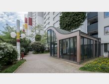 Wohngebiet mit mehrstöckigen Gebäuden und einem eingeschossigen Eingangsbereich aus Glas mit einem gebogenen Dach. Im Vordergrund befinden sich Pflasterwege, blühende Sträucher, eine Laterne und Grünflächen mit Löwenzahn.