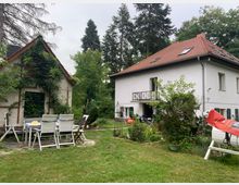 Ein Garten mit viel Grün, einem kleinen weiß gestrichenen Nebengebäude mit Kletterpflanzen und einem weißen Wohnhaus mit rotem Satteldach im Hintergrund. Im Vordergrund steht ein Gartentisch mit weißen Stühlen auf einer gepflasterten Fläche.