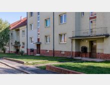 Mehrfamilienhaus aus den 1920er Jahren mit hell beigem Putz und roten Klinkerdetails am unteren Fassadenbereich. Vor dem Gebäude befinden sich kleine grüne Vorgärten, ein gepflasterter Gehweg und niedrige Backsteinumrandungen.