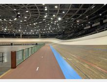 Innenansicht einer Velodrom-Halle mit einer ovalen Holz-Rennbahn und umliegenden Sitztribünen. Die Decke ist mit einem komplexen Metallgerüst und zahlreichen Beleuchtungen ausgestattet.