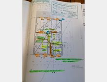 Grundrisszeichnung eines Gebäudes mit Markierungen in verschiedenen Farben (grün, orange, gelb und blau), die räumliche Funktionen, Wände und zusätzliche Anmerkungen kennzeichnen. Oben rechts sind Prüfstempel und technische Informationen zu den Bauvorschriften sichtbar.