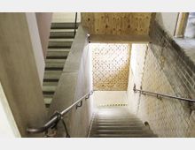 Ansicht einer Betontreppe mit Metallgeländern, die zu einem Bereich aus gelblichen Backsteinwänden führt. Der Raum ist roh und unfertig, mit einem Holzzaun im oberen Bereich und einer deutlichen Trennung zwischen den Treppenabsätzen.