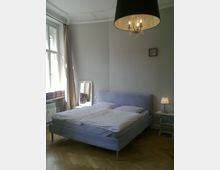 Ein Schlafzimmer mit einem Doppelbett, hellem Holzfußboden und hohen weißen Wänden. Links sind ein Fenster mit beige Vorhängen und ein Heizkörper, oben hängt ein schwarzer Kronleuchter, und rechts ein kleiner Nachttisch mit Tischlampe.