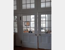 Weiße Doppeltüren mit Glasfenstern führen in einen hellen Raum, in dem ein Tisch mit Stühlen, ein Sessel und ein kleiner Beistelltisch stehen. Durch die Fenster sind weitere große Fenster sichtbar, die den Raum mit Tageslicht durchfluten.