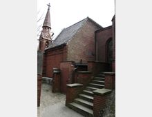 Backsteinbau einer Kirche mit einem steilen Satteldach und einem schlanken Turm mit Kreuz. Im Vordergrund sind eine Treppe aus Stein, mehrere rote Backsteinmauern und ein Seiteneingang zu sehen.
