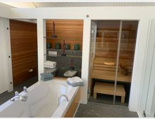 Modernes Badezimmer mit freistehender Badewanne, Glasduschkabine und integrierter Holzsauna. Im Hintergrund sind Regale mit dekorativen Elementen und Handtüchern, sowie Holzvertäfelungen sichtbar.