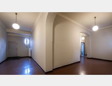 Flur mit glatten, braunen Böden, cremefarbenen Wänden und abgerundeten Türbögen. Deckenlampen mit runden, leuchtenden Kugelleuchten sowie ein kleines, rundes Fenster und eine geschlossene Metalltür sind sichtbar.