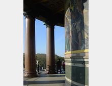 Blick auf eine historische Säulenhalle mit braunen Marmorsäulen, verziertem Gesims und einem Mosaik mit goldenen Details auf einer der Säulen. Im Hintergrund sind Menschen, Bäume und ein klarer blauer Himmel zu sehen.