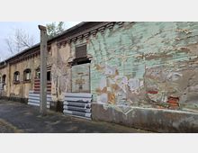Verwitterte Außenwand eines alten Industrie- oder Lagergebäudes mit bröckelndem Putz, freiliegendem Mauerwerk und verblassten Graffiti. Entlang der Fassade stehen ein Betonmast und gestapelte Dämmplatten; kleine Fensteröffnungen und eine Lüftungsöffnung sind zu sehen.
