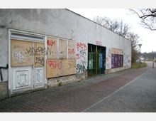 Eine verlassene Gebäudeaußenwand mit verschlossenen und teils verbarrikadierten Türen und Fenstern, bedeckt von zahlreichen Graffiti. Der angrenzende Gehweg ist aus Pflastersteinen, und im Hintergrund sind kahle Bäume und eine Straße sichtbar.