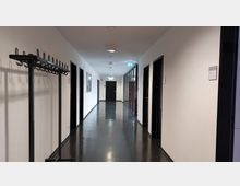 Ein moderner, heller Büroflur mit glänzendem dunklem Fußboden, weißen Wänden und mehreren schwarzen Türen auf beiden Seiten. An der linken Wand steht ein freistehender Kleiderständer, und am Ende des Flurs befindet sich eine geschlossene Tür unter einem Notausgangsschild.