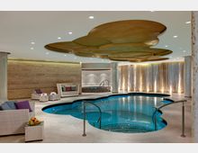 Beleuchteter Innenbereich eines Spas mit einem geschwungenen Pool, weiß gefliesten Böden und hellen Wänden. Im Hintergrund befinden sich Liegen mit weißen Polstern, eine gelbliche Holzverkleidung sowie lichtdurchlässige Vorhänge; die Decke ist mit goldfarbenen, wellenförmigen Elementen gestaltet.