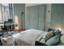 Ein multifunktionaler Raum mit einem Bett, mehreren Kissen und einer grauen Tagesdecke im Vordergrund. Im Hintergrund stehen ein Schreibtisch mit einem Bürostuhl, Studiomonitoren, ein Mikrofonständer sowie Akustikpaneele an den Wänden; das Fenster lässt Tageslicht herein.