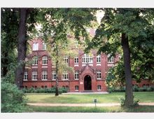 Backsteinbau im neugotischen Stil mit großen Fenstern und einem mittigen Rundbogenportal, umgeben von Bäumen und Grünflächen in einem parkähnlichen Umfeld.