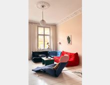 Wohnzimmer mit hellen Wänden, großen Fenstern und Ornamentleisten an der Decke. Im Raum befinden sich farbige Polstermöbel in Rot, Blau und Schwarz, ein niedriger Couchtisch und eine moderne Hängelampe; lichtdurchflutete Atmosphäre mit Vorhängen und Wanddekoration.