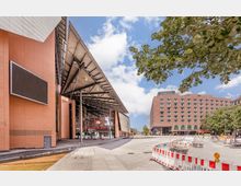 Moderne Gebäudekomplexe mit orangefarbenen und beigefarbenen Fassaden, darunter die „Spielbank Berlin“ mit einem Glasdach auf Metallstützen. Im Vordergrund eine Baustelle mit Absperrungen und im Hintergrund ein mehrstöckiges Hotelgebäude sowie Bäume.