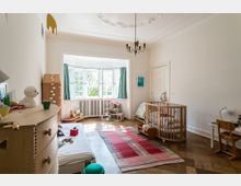 Helles Kinderzimmer mit einem großen Erkerfenster, das von grünen Vorhängen eingerahmt ist. Der Raum enthält ein Babybett, ein kleines Bett, eine bunte Spielzeugaufbewahrung, einen Schreibtisch und eine Kommode, auf einem Holzparkettboden mit einem roten Teppich.