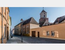 Straßenszene in einer historischen Kleinstadt mit gepflastertem Gehweg, Wohnhäusern aus verschiedenfarbigem Putz und einer Kirche mit Glockenturm im Hintergrund. Die Architektur zeigt Backstein- und Fachwerkelemente unter klarem Himmel.