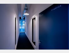 Langer, schmaler Flur mit weißen Wänden und einer dunklen Tür im Vordergrund, auf der „Friedrich“ und „Auguste“ steht. Am Ende des Flurs befindet sich ein großes, blau getöntes Fenster, das Tageslicht hereinlässt, sowie an der Decke moderne Hängelampen.