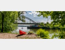 Eine Uferlandschaft mit einer Brücke im Hintergrund, unter der ein Fluss oder See verläuft. Im Vordergrund liegt ein rotes Kanu am sandigen Ufer, umgeben von dichter Vegetation, während eine Person auf einem Stand-Up-Paddleboard auf dem Wasser zu sehen ist.
