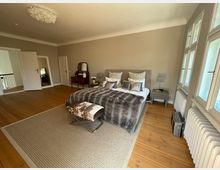 Großes Schlafzimmer mit einem Doppelbett, das mit gemusterten Kissen und einer Felldecke dekoriert ist. Der Raum hat Holzboden, einen cremefarbenen Teppich, einen Schminktisch mit Spiegel, mehrere Fenster mit weißem Rahmen auf der rechten Seite und helle Wände.