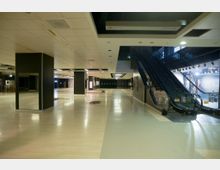 Leerer Innenbereich eines Einkaufszentrums mit hellem Boden und einer Rolltreppe rechts, die in ein Obergeschoss führt. Dunkle Wände und Säulen dominieren den Raum, der von Deckenbeleuchtung schwach erleuchtet ist.