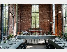 Ein großer, hoher Raum mit unverputzten Ziegelwänden und großen, bodentiefen Fenstern. Die Einrichtung besteht aus festlich gedeckten Tischen mit weißen Tischdecken, grünen Pflanzenarrangements und Kerzen, passend für eine Veranstaltung oder Feier.