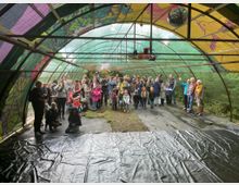 Eine überdachte Veranstaltungsfläche mit einem bunten, bemalten Dach aus transparenten und gemusterten Materialien. Unter der Überdachung steht eine größere Gruppe von Menschen, darunter Erwachsene und Kinder, vor einer mit schwarzer Plane bedeckten Bühne; im Hintergrund befinden sich Bäume.