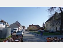 Straßenszene in einer Kleinstadt mit Kopfsteinpflaster, flankiert von Wohngebäuden und kleinen Läden. Im Hintergrund ist ein Kirchturm zu sehen, während im Vordergrund Schuhe auf einem Marktstand ausgestellt sind.