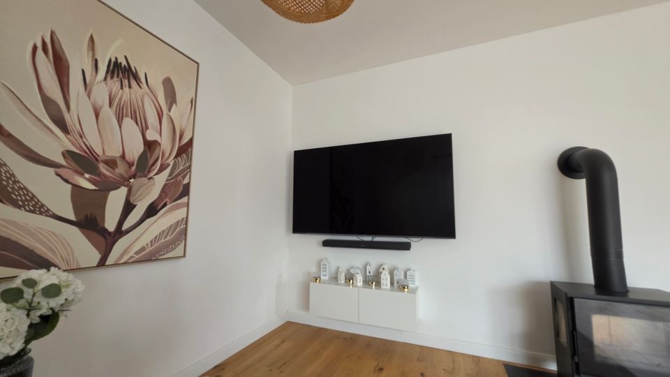 Helle Wohnzimmerecke mit weiß gestrichenen Wänden, Holzboden und einem an der Wand montierten Flachbildfernseher über einem schwebenden Lowboard mit kleinen Deko-Häusern. Links hängt ein großes Blumenbild, rechts steht ein schwarzer Kaminofen mit Ofenrohr; an der Decke hängt eine geflochtene Rattanlampe.