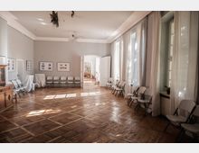 Ein heller Veranstaltungsraum mit Holzboden und weißen Wänden, gesäumt von gefalteten Stühlen entlang der Wand. Große Fenster mit weißen Vorhängen und eine offene Tür, die in einen weiteren hellen Raum führt, sind sichtbar.