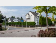 Ein Wohngebiet mit Einfamilienhäusern, modernen Giebeldächern und weißen Fassaden. Die Straße ist von Bäumen, Hecken und einem Gehweg gesäumt, und ein Straßenschild markiert die Kreuzung „Hochkalterweg“.