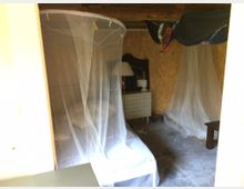 Ein einfach eingerichteter Raum mit Holzwänden, einem Bett mit einem weißen Moskitonetz, einer kleinen Kommode, einem Spiegel und einer weiteren Sitzgelegenheit. Der Boden besteht aus Beton, und der Raum wirkt minimal dekoriert.