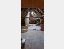 Ein unbehandelter Dachboden mit freiliegenden Holzbalken, einer Betonstütze und schräger Holzverkleidung. Der Boden besteht aus rohem Beton, und der Raum ist mit Kartons und vereinzelten Gegenständen unaufgeräumt.