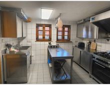 Moderne Großküche mit weißen Fliesenwänden, einem langen Edelstahltisch in der Mitte, mehreren Arbeitsflächen, einem großen Gasherd mit Dunstabzugshaube und Holzrahmenfenstern an der Rückwand. Die Einrichtung umfasst Spüle, Toaster und Kochutensilien.