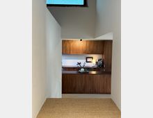View down a narrow white-walled hallway into a modern kitchen with walnut wood cabinets and a matching island, lit by a small overhead spotlight. A high rectangular window near the ceiling brings in daylight above the kitchen area.