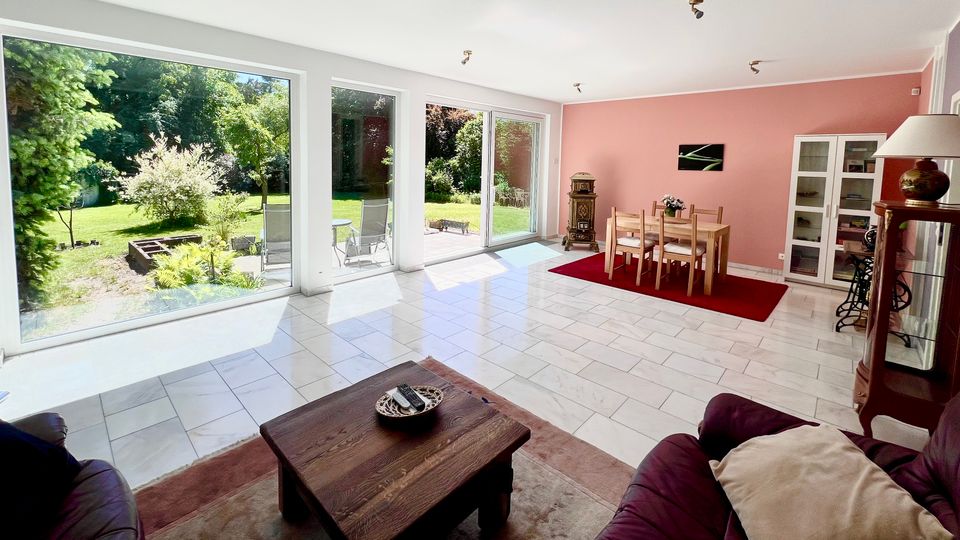 Heller Wohn- und Essbereich mit großen bodentiefen Fenstern und Glasschiebetür zur Terrasse und in einen grünen Garten. Fliesenboden, rosa Akzentwand sowie Esstisch auf rotem Teppich und Wohnmöbel im Vordergrund.