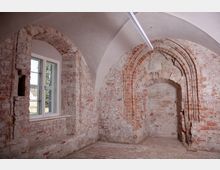 Innenraum eines historischen Gebäudes mit freigelegten Backsteinmauern, gotischen Spitzbogen-Elementen und einem weiß gestrichenen Gewölbedeckenabschluss; links ein modernes Fenster mit Blick ins Freie.