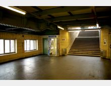 Unterirdischer Bahnhofszugang mit gelb gefliesten Wänden, einer Treppe, die nach oben führt, und einer Fotokabine auf der rechten Seite. Die Decke besteht aus Metallträgern, und die Beleuchtung erfolgt durch lange Neonröhren.
