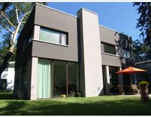 Modernes zweistöckiges Wohnhaus mit glatter, grauer Putzfassade und großen Glasfronten. Vor dem Haus befindet sich eine Terrasse mit einem orangefarbenen Sonnenschirm, Sitzmöbeln und einem gepflegten Rasen, umgeben von hohen Bäumen.