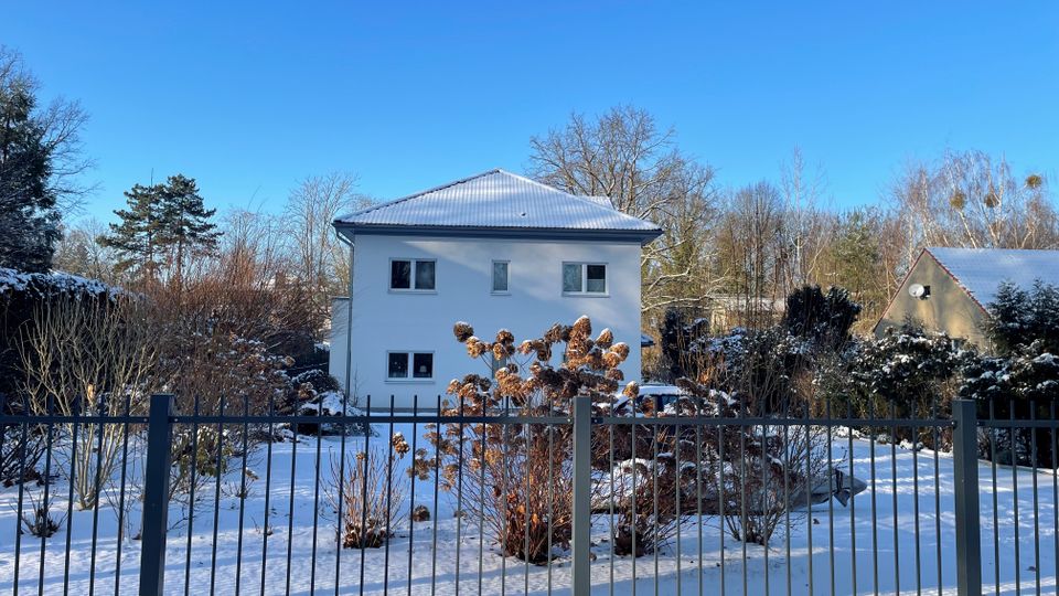 Blick auf ein freistehendes, zweigeschossiges Wohnhaus mit weißer Fassade und leicht verschneitem Walmdach in einem winterlichen Garten. Im Vordergrund verläuft ein dunkler Metallzaun, dahinter liegen schneebedeckte Beete und Sträucher sowie kahle Bäume unter blauem Himmel.