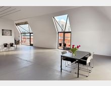 Heller, moderner Innenraum mit großen, schrägen Fenstern, durch die Tageslicht einfällt. Im Vordergrund steht ein Tisch mit Stühlen und einem Blumenstrauß, im Hintergrund sind weitere Stühle und ein Balkon mit Blick auf ein Ziegelgebäude sichtbar.