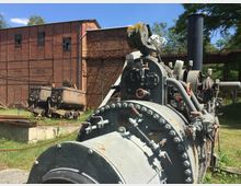 Außenansicht eines historischen Industriestandorts mit einer rostigen Dampfmaschine im Vordergrund, einem alten Förderwagen und einem Backsteingebäude im Hintergrund. Umgeben von Grünflächen und einzelnen Bäumen.