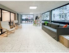 Heller Aufenthaltsbereich in einem Gebäude mit großflächigen Fensterfronten, gefliestem Boden und Deckenleuchten. Links stehen zwei helle Sessel vor Glaswänden, rechts ein graues Sofa; im Hintergrund ein kleiner Stehtisch mit Hockern und Blick in einen Hofbereich.