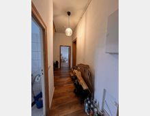 Enger Flur in einer Wohnung mit Holzfußboden, weiß gestrichenen Wänden und einer hängenden Deckenleuchte. Rechts sind Kartons und Glasflaschen gestapelt, links führt eine Tür zu einem Badezimmer mit weißen Fliesen.