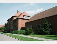 Backsteingebäude im klassischen Stil mit großen Sprossenfenstern und roten Ziegeldächern, umgeben von Grünflächen, Gehwegen und einzelnen Bäumen. Der Himmel ist klar und blau.