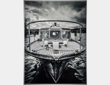 Schwarz-Weiß-Aufnahme eines Bootes auf einem See, aufgenommen von der Vorderseite. Das Boot hat ein offenes Deck mit Holzplanken und ein geschlossenes Cockpit mit großen Fenstern. Im Hintergrund sind bewölkter Himmel und bewaldete Ufer erkennbar.