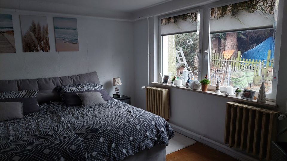 Schlafzimmer mit großem Bett in grauer Bettwäsche, weißen Wänden und drei Wandbildern über dem Kopfteil. Rechts eine breite Fensterfront mit Fensterbank voller kleiner Dekorationen und Blick in einen Garten; darunter stehen zwei Heizkörper.